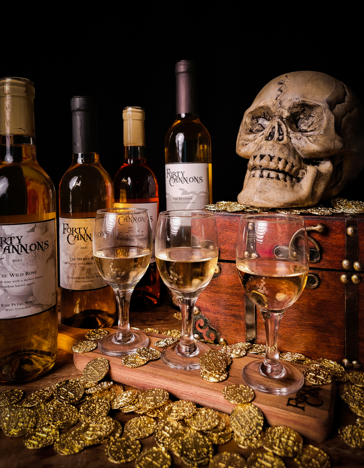 Three small wine glasses with wine bottles in the background, gold coins on the table, and a skull overlooking the setting