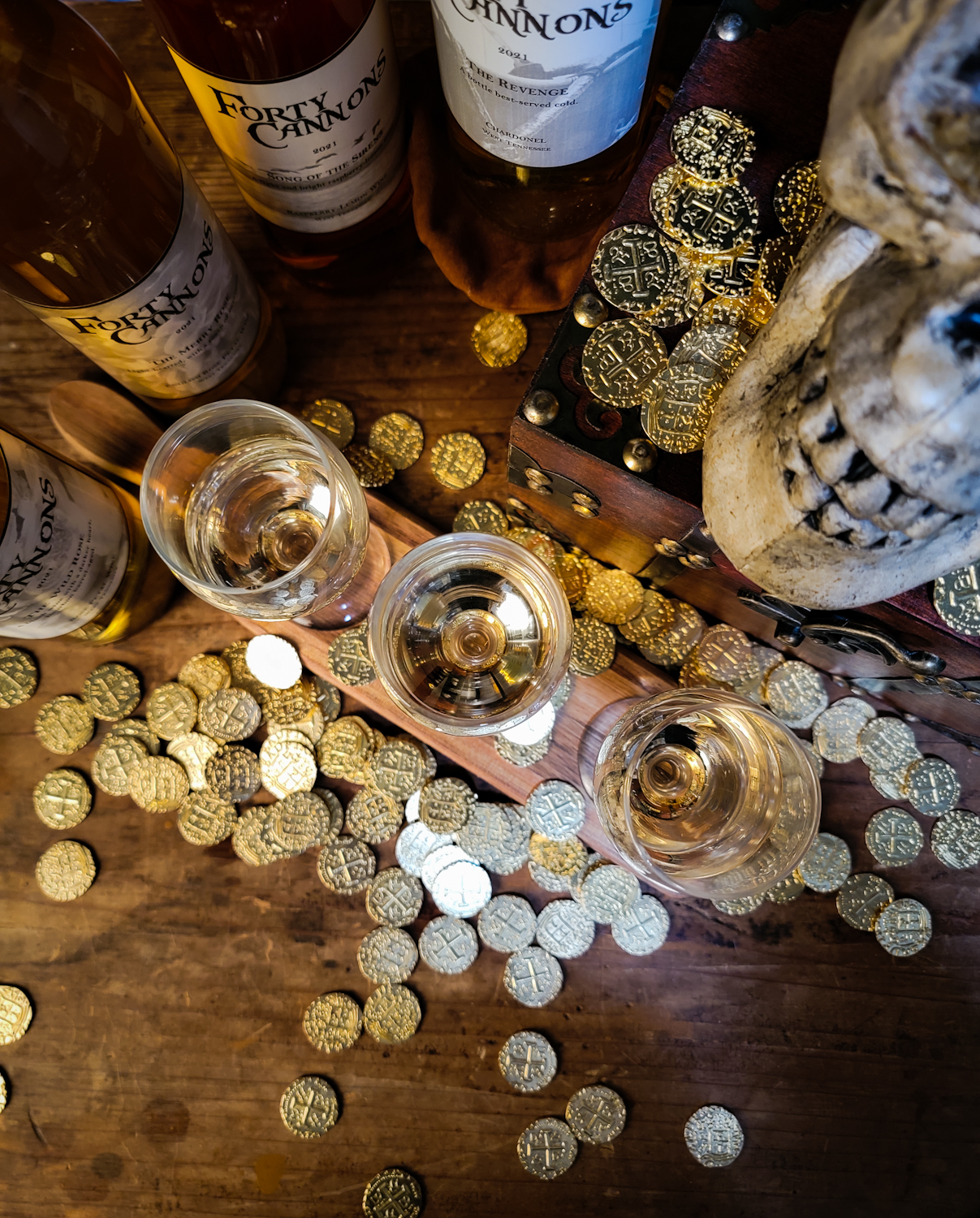 Overhead view of three small wine glasses with wine bottles in the background, gold coins on the table, and a skull overlooking the setting
