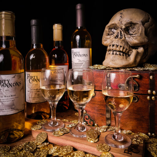 Three small wine glasses with wine bottles in the background, gold coins on the table, and a skull overlooking the setting